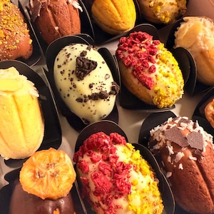 May include: An assortment of colorful, gourmet madeleines in a box. The madeleines are decorated with various toppings, including chocolate, coconut flakes, dried fruit, and nuts. The treats are arranged in individual black paper cups.