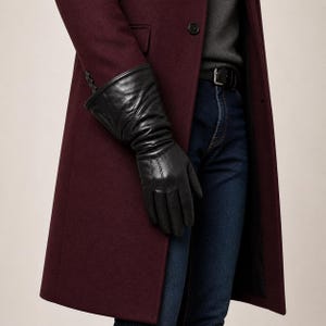 May include: Black leather gloves with a pleated design on the wrist. The gloves are worn with a burgundy coat and dark blue jeans. The coat has a single button closure.