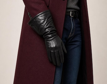 May include: Black leather gloves with a pleated design on the wrist. The gloves are worn with a burgundy coat and dark blue jeans. The coat has a single button closure.