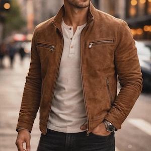 May include: A tan suede jacket with a stand-up collar, a full-length front zip, and two horizontal zipped chest pockets. The jacket is worn over a cream-coloured henley shirt with a buttoned placket, paired with dark denim jeans.