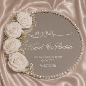 May include: A circular mirror tray decorated with a border of white pearls and a cluster of four white roses with baby's breath. The mirror features white text: Bismillah, Navid & Shaista, And We created you in pairs, Qur'an (78:8), and 26/07/2026.