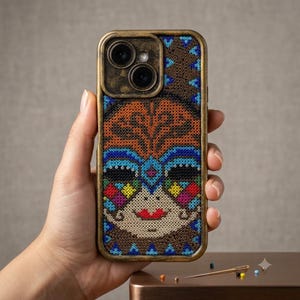 May include: A smartphone case featuring a detailed beadwork design of a stylised face. The pattern uses orange, blue, black, red, yellow, and cream-coloured beads. The case has a metallic bronze-toned frame surrounding the camera lens cutout.
