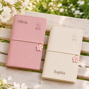 Può includere: Due taccuini rettangolari, uno rosa polvere e uno color crema, ciascuno dotato di un elastico verticale con un ciondolo a forma di fiore di ciliegio rosa. Il taccuino rosa è inciso con il nome Olivia, quello crema con Sophia.
