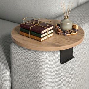 May include: A round wooden side table with a black metal bracket, resting on a gray upholstered sofa. The table holds three stacked books, gold-rimmed glasses, a small vase with reeds, and decorative items like cinnamon sticks and dried fruit.