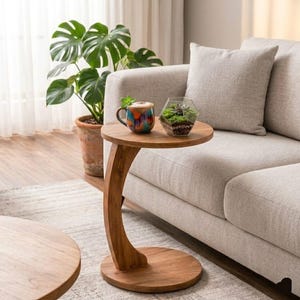 May include: A wooden side table with a curved leg and round top. A colorful mug and a geometric terrarium sit on the table. A potted plant and a beige sofa are in the background.