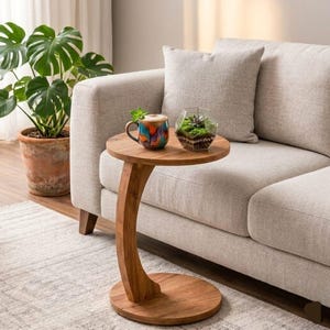 Puede incluir: Una mesa auxiliar de madera con tapa y base redondas, unidas por una pata curva. Una taza colorida y un pequeño terrario están sobre la mesa. La mesa está junto a un sofá de color claro y una planta en maceta.
