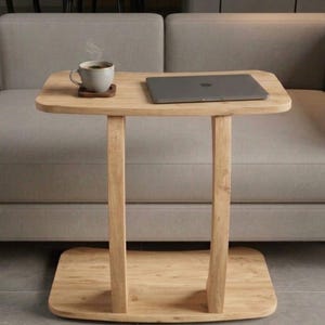 Puede incluir: Una mesa auxiliar de madera clara con una parte superior rectangular y un estante inferior. Una computadora portátil y una taza de café están sobre la mesa. La mesa tiene un diseño minimalista y está colocada frente a un sofá gris.