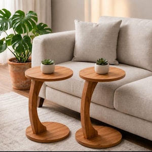 May include: Two wooden side tables with a round top and curved leg. Each table has a small white pot with a succulent plant on top. The tables are made of light brown wood and sit on a light-colored rug.