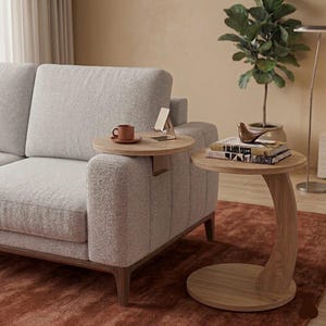 May include: A light grey sofa with a wooden frame and two attached side tables. One table holds a brown cup and a phone stand, the other holds books and a decorative bird. The tables are made of light wood and sit on a red rug.
