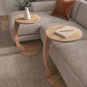 May include: Two wooden side tables with a curved leg design. The round tabletops are made of light-coloured wood, and one holds a white vase. The tables are placed next to a grey sofa with a brown cushion.