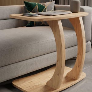 Op de afbeelding: Een licht houten bijzettafel met een rechthoekig blad en gebogen poten. De tafel is ontworpen om onder een bank te schuiven. Een laptop, telefoon en luidspreker staan op het tafelblad.