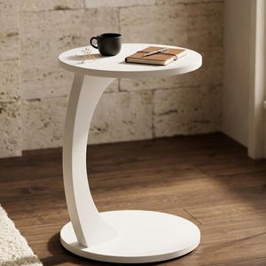 Puede incluir: Una mesa auxiliar blanca en forma de C con una superficie redonda. Una taza de café negra y un pequeño cuaderno descansan sobre la mesa. La mesa tiene una pata curva y una base circular. El fondo presenta una pared texturizada y un suelo de madera.