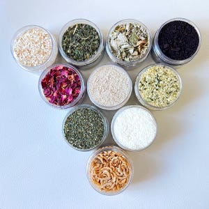 May include: Ten clear circular containers arranged in a triangle formation, each filled with different dry ingredients including dried rose petals, green herbs, white powder, shredded coconut, dark coffee grounds, and small dried shrimp.