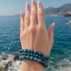 Könnte beinhalten: Drei blaue Perlenarmbänder mit silbernen Details sind an einem Handgelenk gestapelt. Die Armbänder bestehen aus runden, gesprenkelten Perlen. Der Hintergrund zeigt ein blaues Meer und einen blauen Himmel.