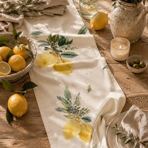 Könnte beinhalten: Ein cremefarbener Tischläufer mit einem Aquarell-Zitronen- und Blattdesign. Der Läufer liegt auf einem Holztisch, zusammen mit Zitronen, Oliven, einer Kerze und einer Vase.