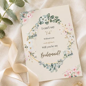 Könnte beinhalten: Eine rechteckige Karte mit cremefarbenem Hintergrund und einem goldenen kreisförmigen Rahmen, der mit Aquarell-Eukalyptusblättern sowie weißen und rosa Blüten verziert ist. Der Text lautet: I can't say "I do" Without you Will you be my Bridesmaid?