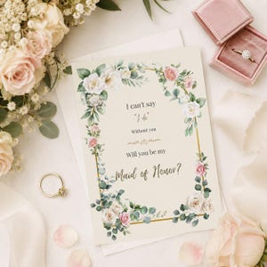 Könnte beinhalten: Eine cremefarbene Karte mit einem goldenen rechteckigen Rahmen, der mit Aquarellrosen und Eukalyptusblättern verziert ist. Der Text lautet: I can't say "I do" Without you Will you be my Maid of Honor? Ein goldener Ring liegt auf der Karte und in einer rosa Schachtel.
