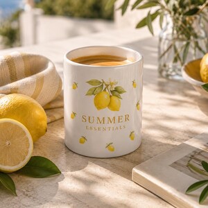 May include: A white ceramic mug filled with coffee, adorned with yellow lemon illustrations and the text "SUMMER ESSENTIALS." The mug sits on a stone surface next to a halved lemon and a folded yellow and white striped cloth.