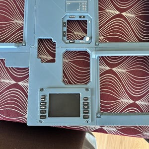 May include: A light blue aircraft control panel with various labeled sections and a digital display. The panel includes text such as "CS-TNJ", "KIKO NERO", and "BUGS". The panel is made of a rigid material and has a matte finish.