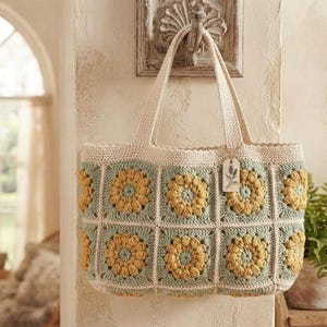 May include: A crochet tote bag featuring a pattern of square motifs with yellow sunflower-like centers and sage green borders. The bag has two cream-colored handles and a small rectangular tag attached to one handle with a floral illustration.