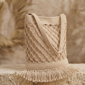 May include: A beige crochet bucket bag featuring a diamond-patterned macrame overlay, a solid inner lining, two sturdy shoulder straps, and a decorative fringe trim along the bottom edge. The bag is constructed with a dense, uniform stitch pattern.