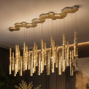 May include: A contemporary chandelier featuring a gold-toned, hexagonal ceiling fixture. Many vertical, cylindrical lights descend from the fixture, each providing a warm, golden illumination. The lights are positioned above a dining table, creating a sophisticated atmosphere.