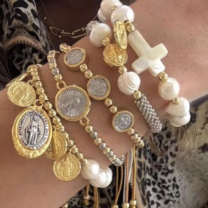 May include: A stack of bracelets featuring white baroque pearls, gold-toned metal beads, and silver-toned circular medallions depicting religious figures. One bracelet includes a white cross-shaped charm, while others feature dangling coin pendants.