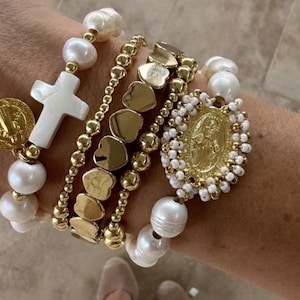 May include: A stack of four bracelets featuring white freshwater pearls, gold-toned metal beads, a white cross charm, a gold-toned circular medal, a row of gold-toned heart-shaped beads, and a central gold-toned oval medal framed by small white beads.