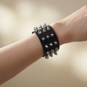 Puede incluir: Brazalete de cuero negro con tachuelas cónicas plateadas. El brazalete ancho se lleva en la muñeca y presenta múltiples filas de tachuelas. El fondo es de un color suave y neutro.