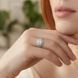 May include: A close-up of a hand wearing a three-piece ring set. The rings are silver-toned with a square, halo-style center stone and smaller stones around the band. The background is blurred, with a focus on the rings.