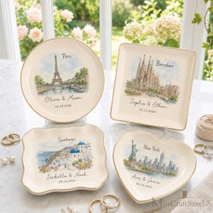May include: Four cream-colored ceramic trinket dishes with gold-toned rims, each featuring a watercolor illustration of a city landmark: Paris, Barcelona, Santorini, and New York. Each dish includes custom names and dates printed in black script.