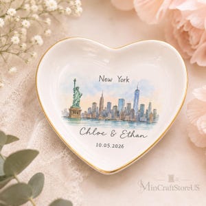 May include: A white heart-shaped ceramic trinket dish with a gold-painted rim. The center features a watercolor illustration of the New York City skyline and the Statue of Liberty, with the text New York, Chloe & Ethan, and 10.05.2026.