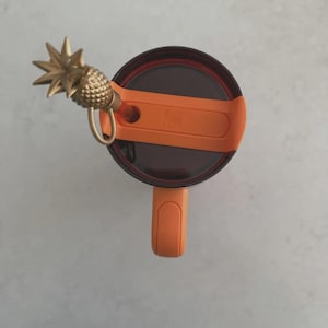 Puede incluir: Un dije de pi&ntilde;a dorado adjunto a una tapa naranja de un vaso. La tapa es marr&oacute;n oscuro y naranja, con un asa. El dije est&aacute; detallado con una superficie texturizada y un lazo para sujetar. El vaso est&aacute; sobre una superficie gris claro.