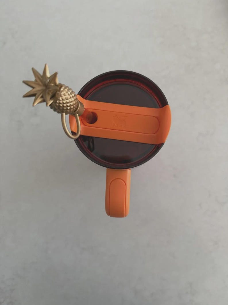 Puede incluir: Un dije de pi&ntilde;a dorado adjunto a una tapa naranja de un vaso. La tapa es marr&oacute;n oscuro y naranja, con un asa. El dije est&aacute; detallado con una superficie texturizada y un lazo para sujetar. El vaso est&aacute; sobre una superficie gris claro.