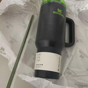 May include: A black Stanley mug with a green lid and handle, featuring the Stanley logo. A green straw is included. The mug is packaged in white paper. The mug has a capacity of 325 ml.