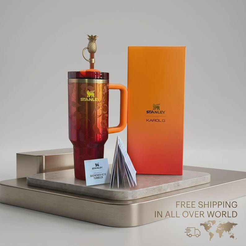 Puede incluir: Un vaso Stanley Quencher H2.0 en degradado rojo y naranja, con un asa naranja a juego. El vaso tiene un tap&oacute;n dorado en forma de pi&ntilde;a y el logotipo de Stanley. Tambi&eacute;n hay una caja con el logotipo de Stanley y "KAROL G".