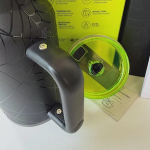 May include: A black insulated water bottle with a handle and a neon green lid. The bottle has a spiderweb design and the lid is partially open. The packaging includes text about features like dishwasher safety and a reusable straw.