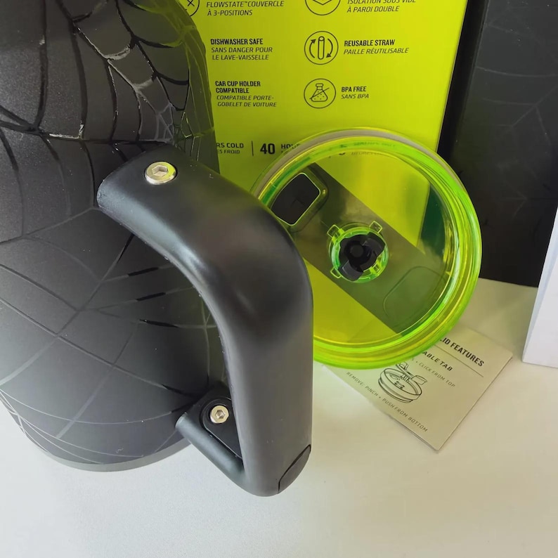 May include: A black insulated water bottle with a handle and a neon green lid. The bottle has a spiderweb design and the lid is partially open. The packaging includes text about features like dishwasher safety and a reusable straw.