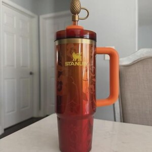 Puede incluir: Un vaso Stanley rojo y naranja con una tapa dorada en forma de pi&ntilde;a. El vaso tiene un asa grande naranja y un dise&ntilde;o floral. El logotipo de Stanley es visible en el lateral.