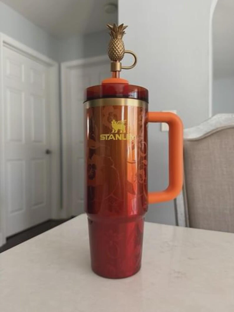 Puede incluir: Un vaso Stanley rojo y naranja con una tapa dorada en forma de pi&ntilde;a. El vaso tiene un asa grande naranja y un dise&ntilde;o floral. El logotipo de Stanley es visible en el lateral.