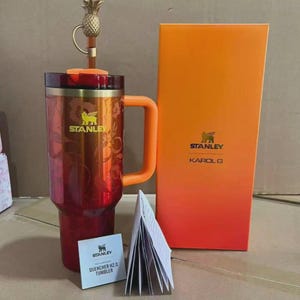 Puede incluir: Un vaso Stanley Quencher H2.0 rojo y naranja con asa, tapa en forma de pi&ntilde;a y dise&ntilde;o floral. El vaso est&aacute; junto a una caja naranja con los logotipos de Stanley y Karol G, y un peque&ntilde;o folleto.