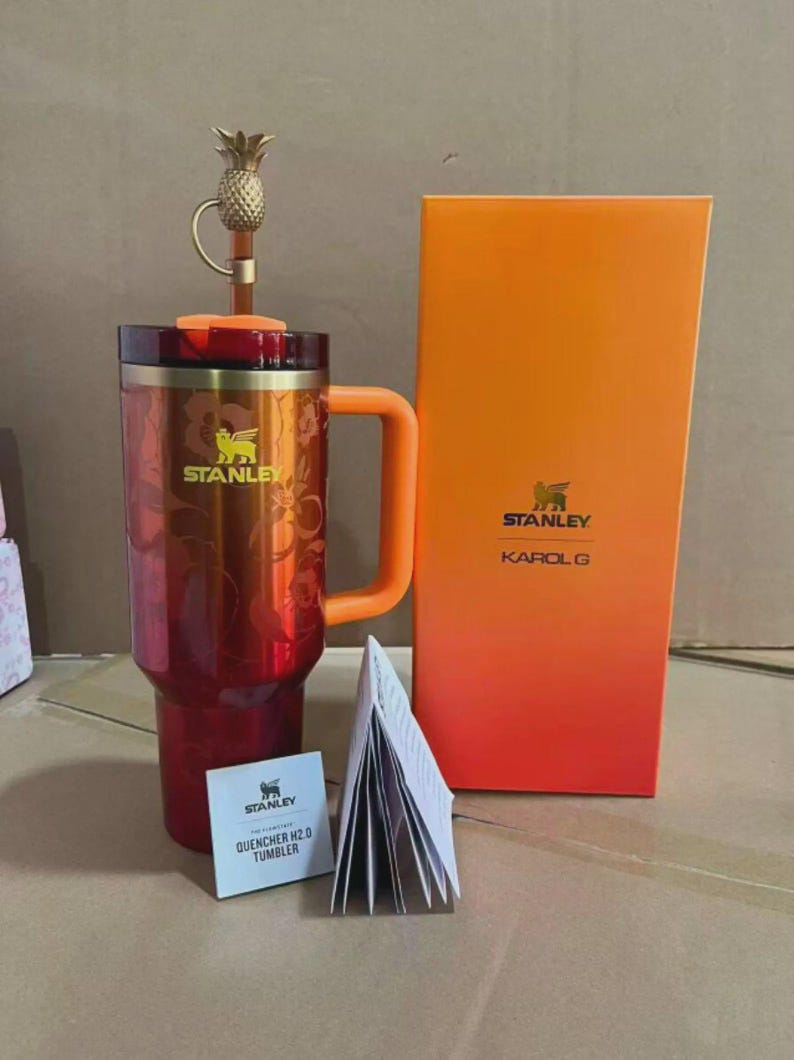 Puede incluir: Un vaso Stanley Quencher H2.0 rojo y naranja con asa, tapa en forma de pi&ntilde;a y dise&ntilde;o floral. El vaso est&aacute; junto a una caja naranja con los logotipos de Stanley y Karol G, y un peque&ntilde;o folleto.