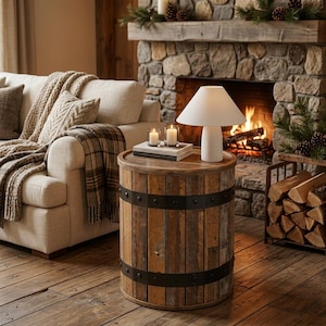 May include: A cylindrical side table constructed from vertical wooden planks with two dark metal bands featuring rivets. The table surface holds two small glass candles and a white lamp with a conical shade, creating a functional furniture piece.
