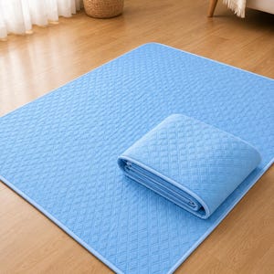 以下が含まれることがあります： 表面全体にキルティング加工されたダイヤモンド模様が施された、長方形のライトブルーのマット。マットには丸みを帯びた角と、同色のライトブルーの布製縁取りが付いています。マットの一部が折り返されており、キルティングの質感が確認できます。