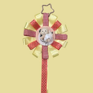 May include: A decorative rosette badge featuring a central circular emblem with a small white lamb figurine. The rosette is constructed from alternating yellow satin and red-and-white gingham ribbons, with a star-shaped top hook and hanging ribbon.