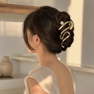 May include: A gold-toned metal hair claw clip with a swirling, abstract design and a single white pearl accent. The clip is secured in dark brown hair styled in an updo, complemented by a small gold hoop earring and a delicate necklace with a pearl pendant.