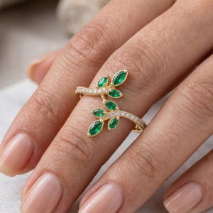 Peut inclure: Une bague en or avec un motif de feuille, ornée de pierres précieuses vert émeraude et de petites pierres claires. La bague s'enroule autour du doigt, créant une esthétique inspirée de la nature.
