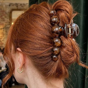 May include: A brown tortoiseshell hair claw clip with a series of round, decorative beads. The clip is holding up auburn hair. A gold hoop earring is visible. The background includes a restaurant setting.