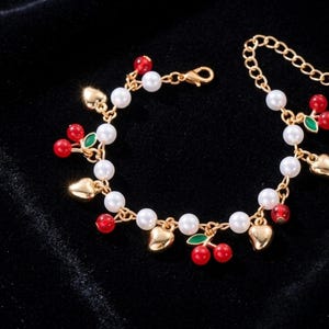 Peut inclure: Bracelet doré orné de fausses perles blanches, de breloques cerises rouges et de breloques cœurs dorés. Le bracelet est doté d'une chaîne d'extension et d'un fermoir mousqueton. Des feuilles vertes ornent les cerises.