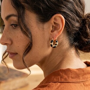 Può includere: Orecchini a cerchio color oro con un design in smalto nero. Gli orecchini sono di piccole dimensioni, di forma circolare e presentano un motivo unico. Il gioiello è indossato sull'orecchio di una persona con i capelli scuri. Un accessorio elegante.